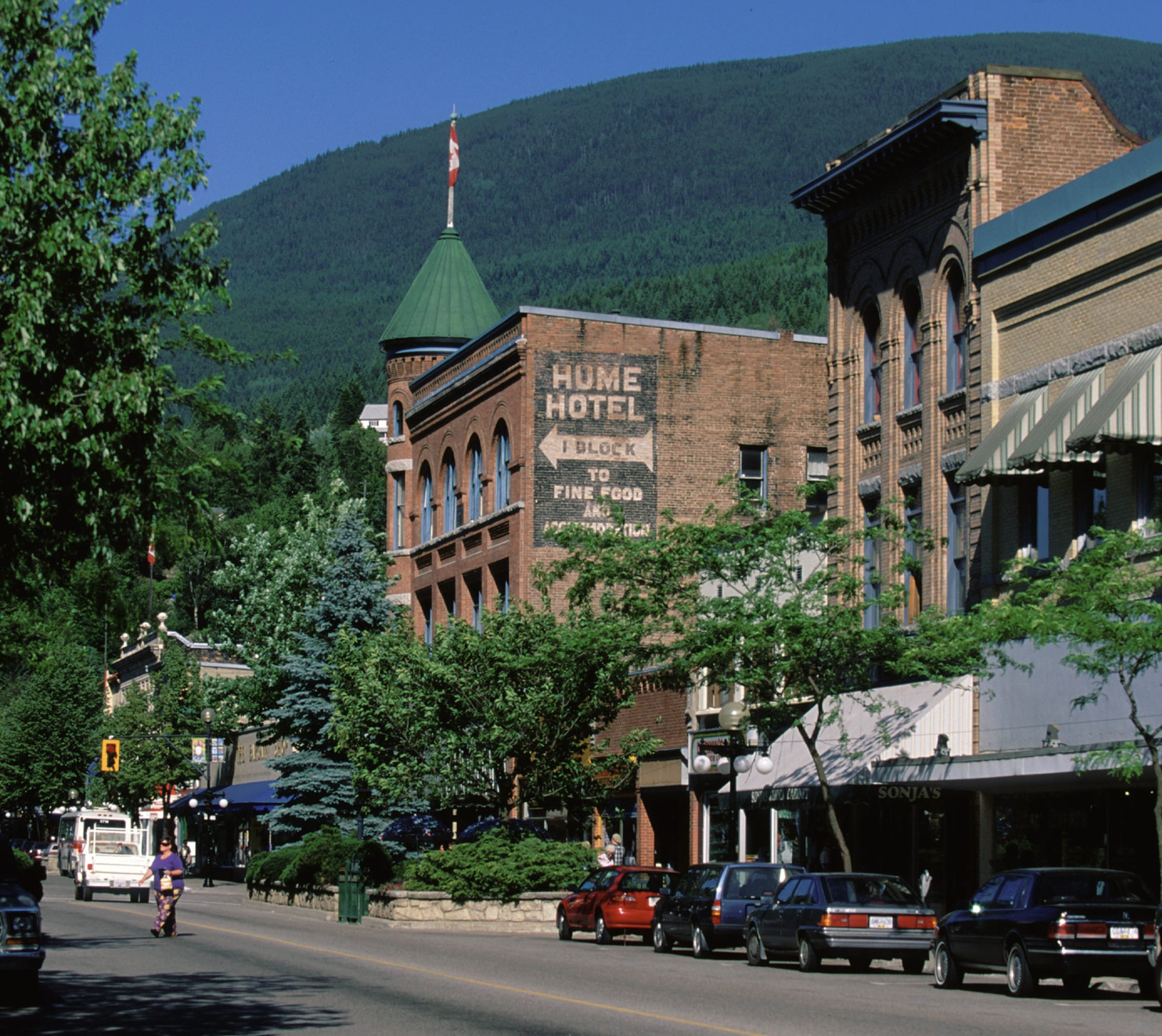Baker Street in Nelson: brick heritage buildings with a green turret and a painted hotel sign, parked cars below and a forested mountain behind.