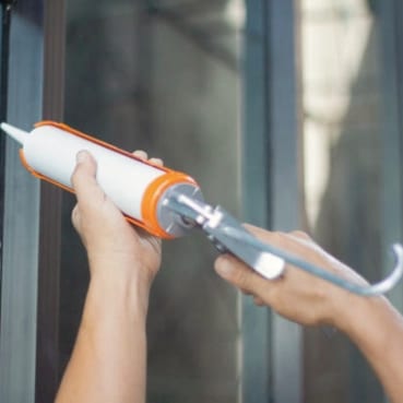 Two hands holding a caulking gun, a tube of sealant loaded, against a glass and metal frame.