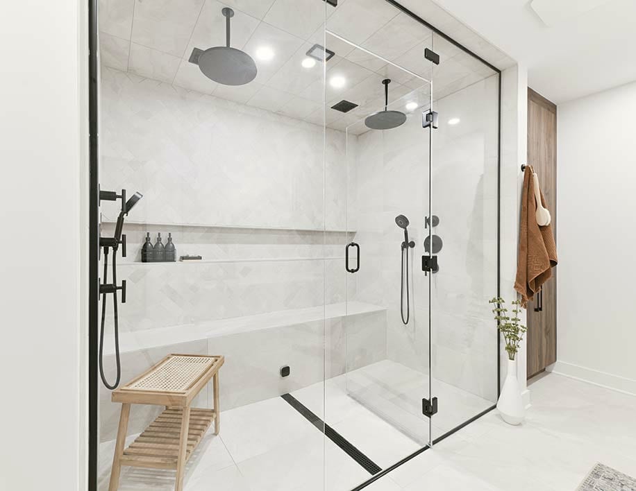 A large walk-in shower behind frameless glass with black hardware, a teak bench inside and two rain heads overhead.